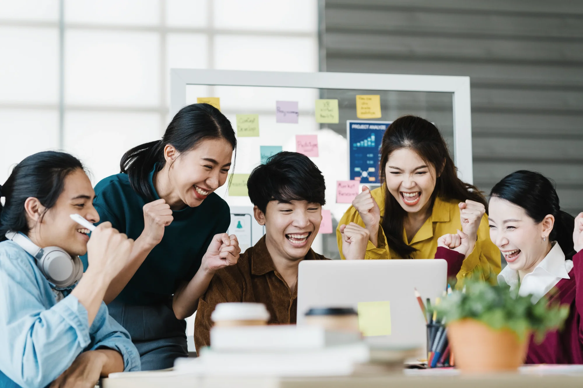 A group of five people employees gather around a laptop, smiling and celebrating. Behind them is a whiteboard with colorful sticky notes. Singapore