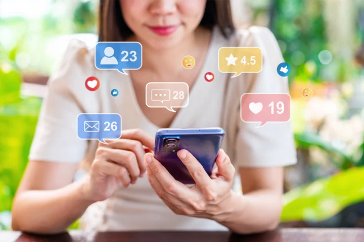 Asian woman uses a smartphone while marketing icons for social media interactions, like comments and likes, appear around her.