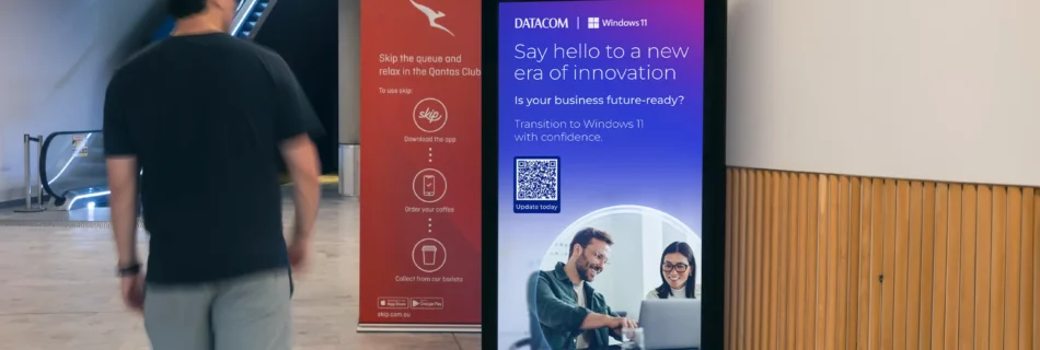 A digital advertising display in a modern indoor Qantas lounges shows a promotional message for Datacom and Windows 11. The ad features a headline reading "Say hello to a new era of innovation," with a subheading, "Is your business future-ready?" It encourages transitioning to Windows 11 with confidence and includes a QR code labeled "Update today." The ad features a photo of two smiling professionals working together on a laptop. Nearby, a smaller red sign promotes Qantas Club with steps to "Skip the queue," referencing an app for ordering coffee. An escalator and wooden wall panels are visible in the background.